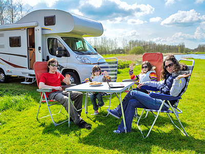 Eine Familie sitzt am Tisch im Freien, neben einem Wohnmobil, und winkt in die Kamera.