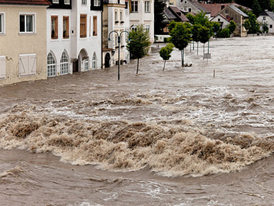 Ein überfluteter Straßenabschnitt mit hohem Wasserstand und Wellen, umgeben von Gebäuden und Bäumen.
